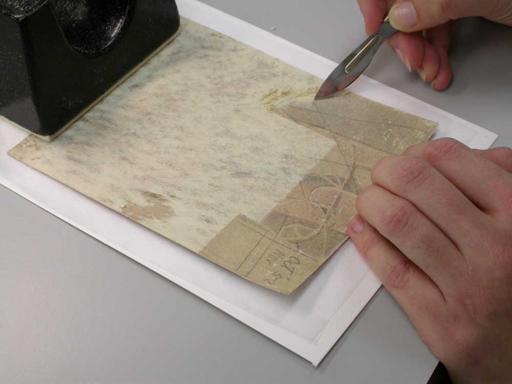 Carefully scraping away the residue of acidic, wood pulp board from the verso of a water-sensitive artwork.